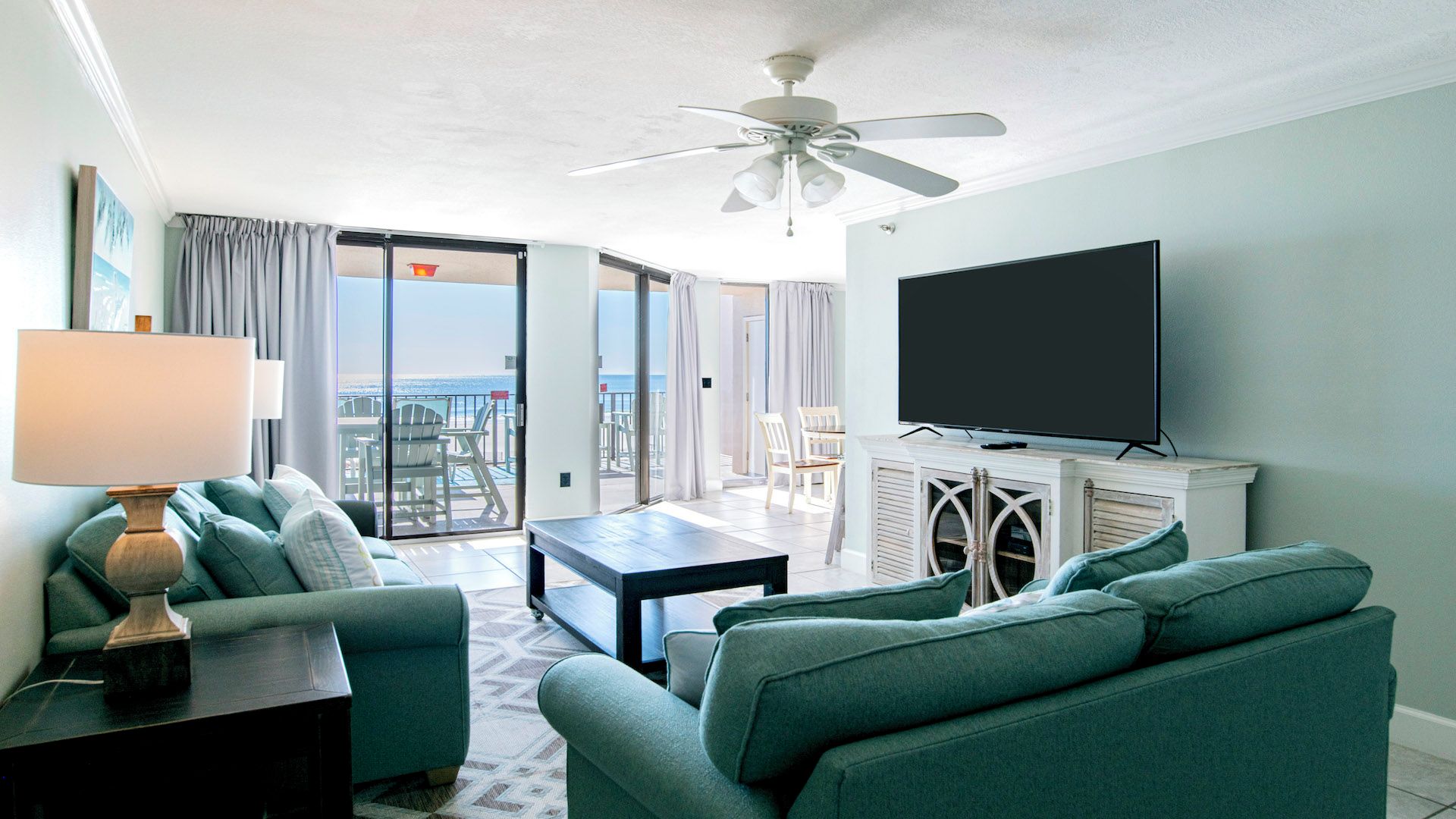 A hotel room with a sofa, a coffee table, a TV, and a ceiling fan. There is a balcony with a view of the ocean.