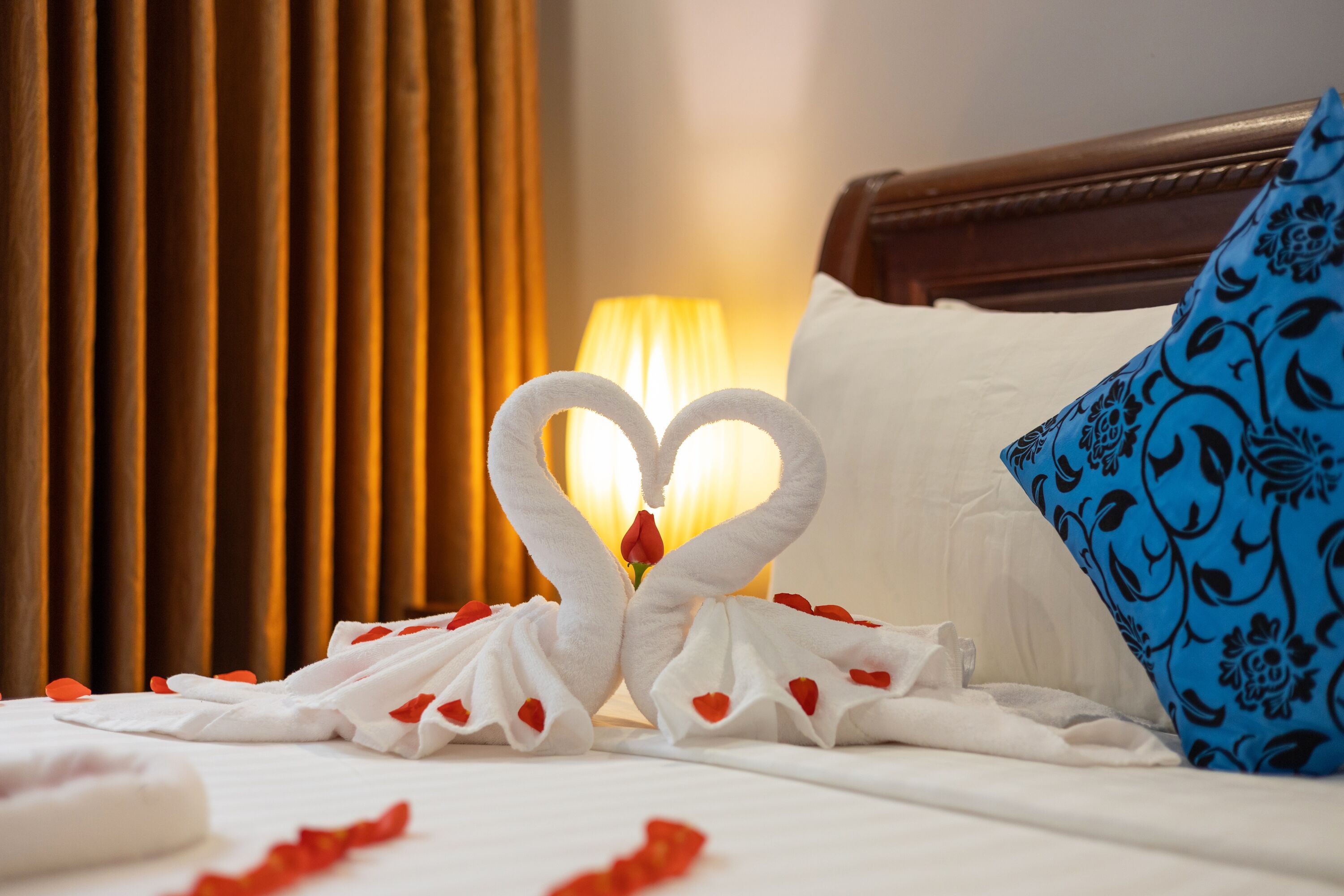 A hotel room with a bed, a heart-shaped towel arrangement, a blue patterned pillow, and a lamp in the background.