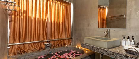 A bathroom with a stone sink, a mirror, and a bathtub with rose petals.
