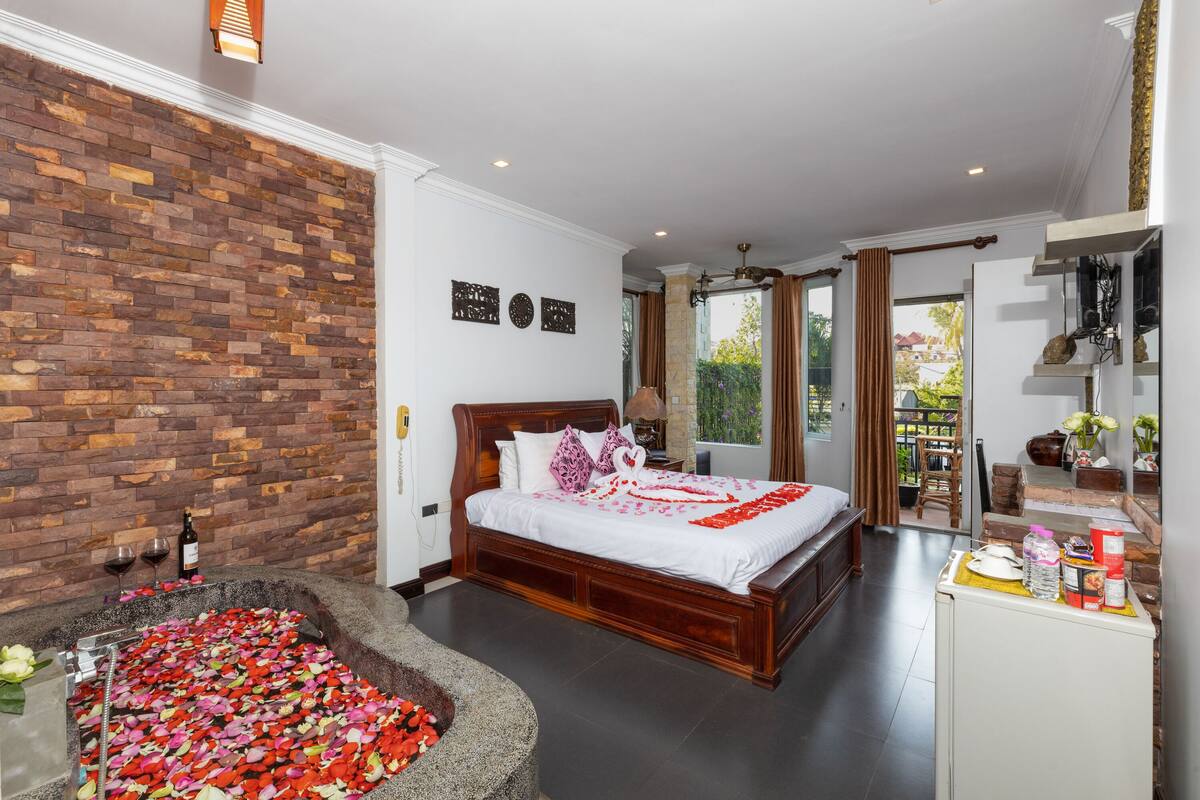 A bedroom with a brick wall, a bed with white bedding, a stone basin with rose petals, and a small kitchenette.