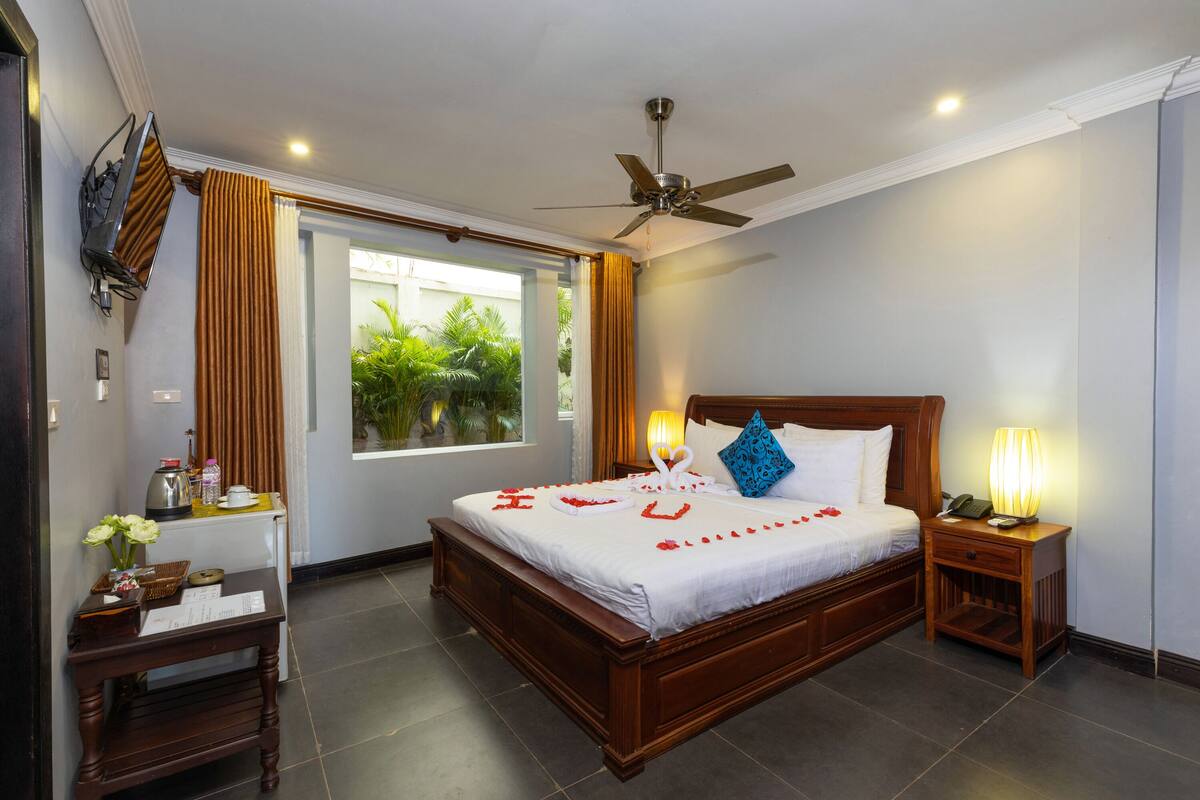 A hotel room with a wooden bed, a nightstand with a lamp, a TV mounted on the wall, and a window with curtains.