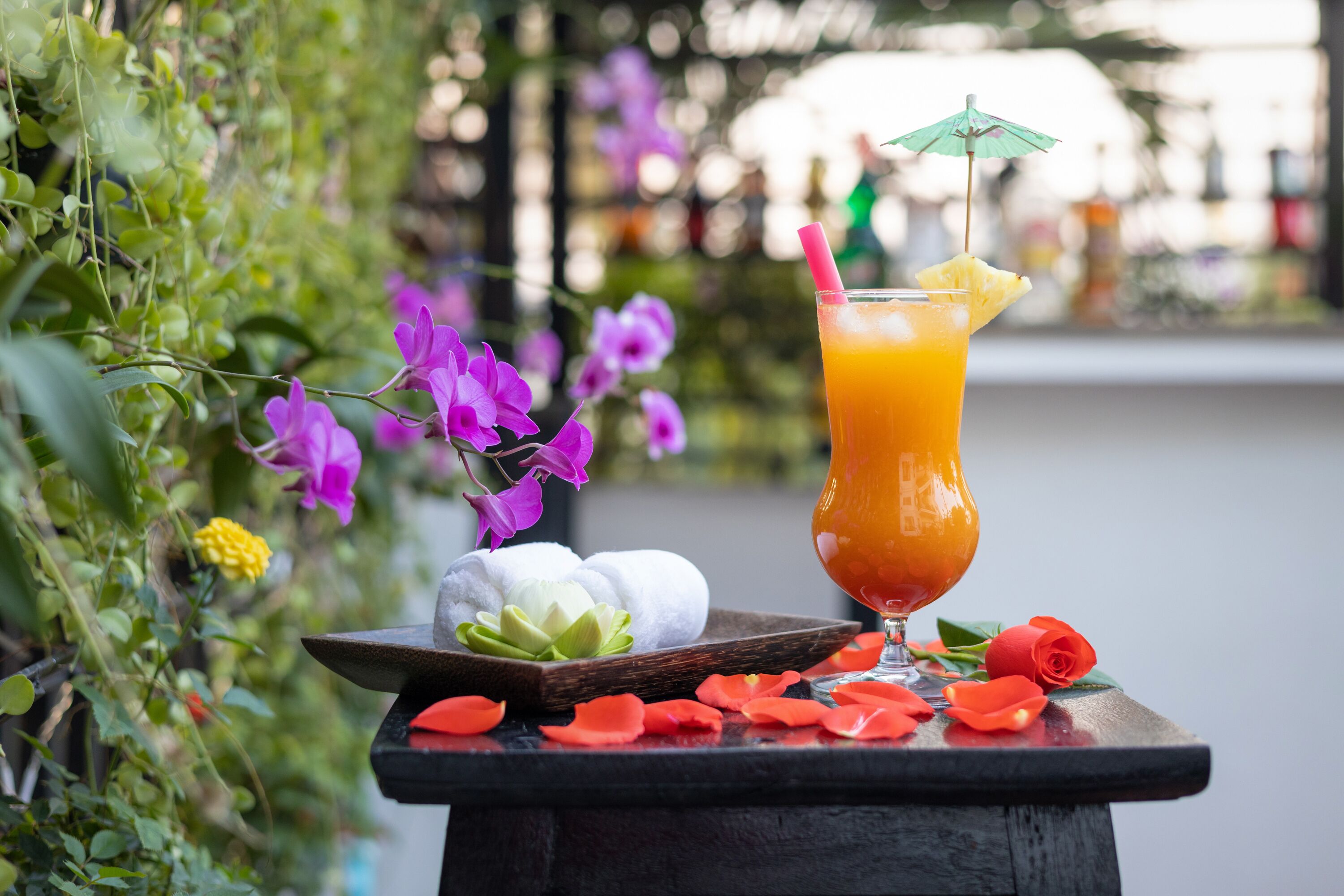 A glass of orange juice with a straw and a pineapple slice on a table with a wooden tray containing a white lotus flower and a red rose.
