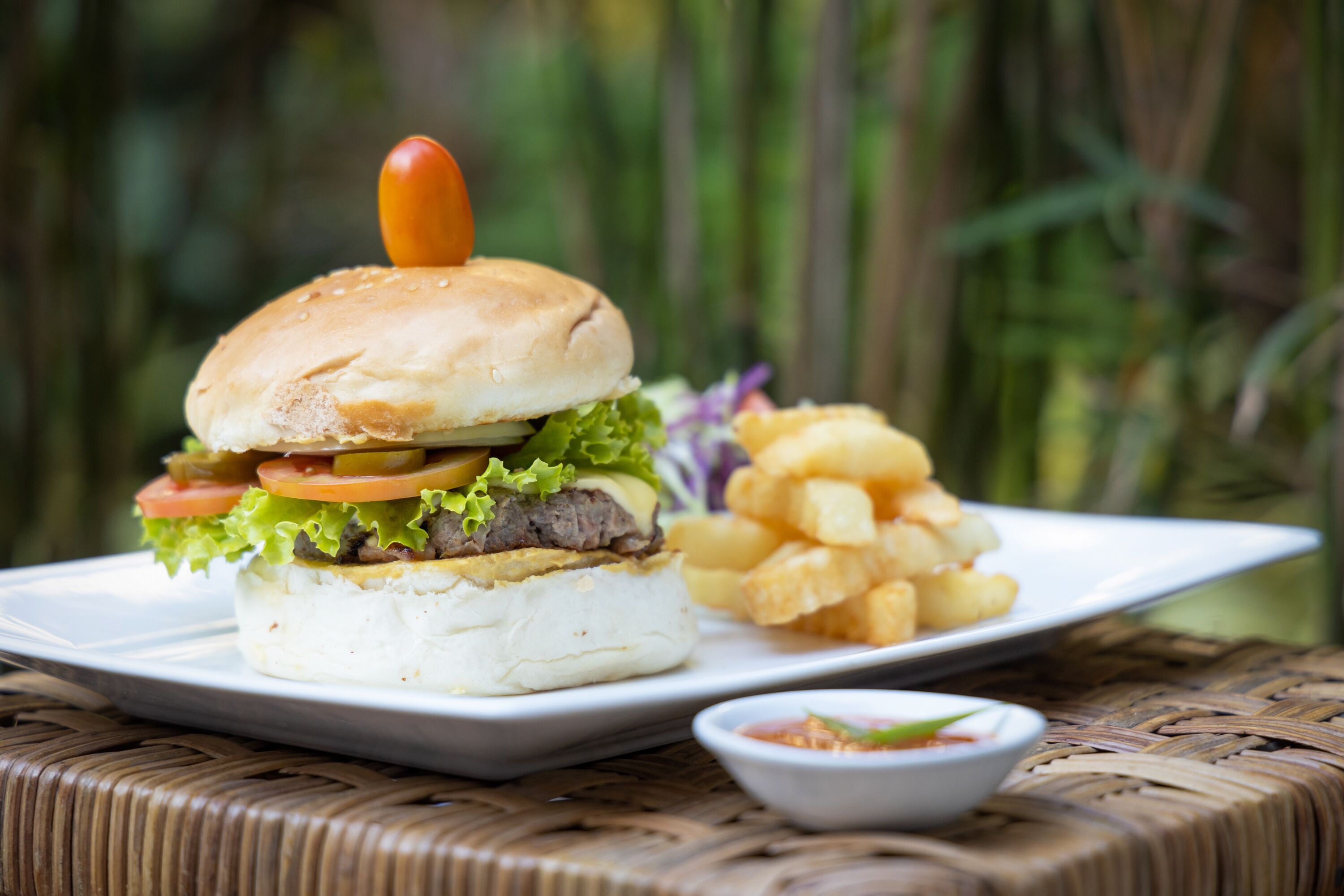 Un sandwich composé d’un pain, de laitue, de tomates et d’une galette de viande, servi avec des frites et une petite portion de sauce dans une assiette blanche.