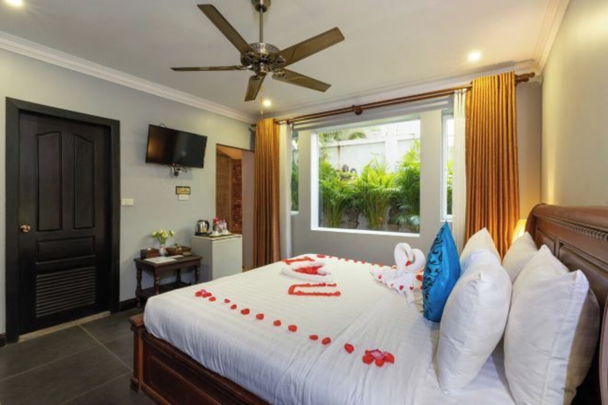 A hotel room with a bed, a ceiling fan, a TV mounted on the wall, and a window with curtains.