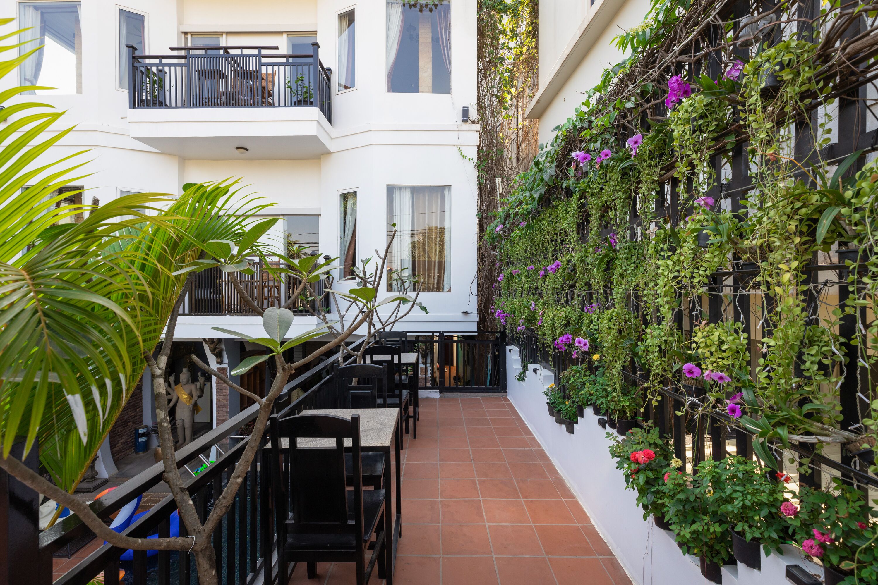 A narrow courtyard with a dining area, white walls, and a balcony with plants and flowers.