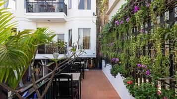 A narrow courtyard with a dining area, white walls, and a balcony with plants and flowers.