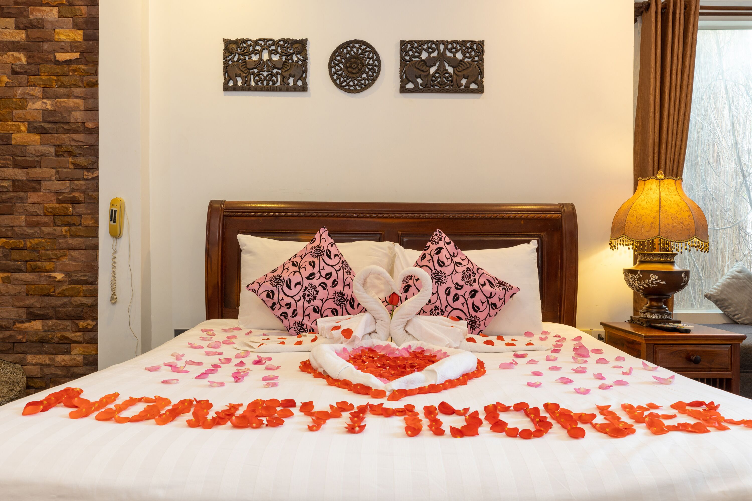 A bed with rose petals and a heart-shaped towel arrangement, a brick wall, and decorative wall art.