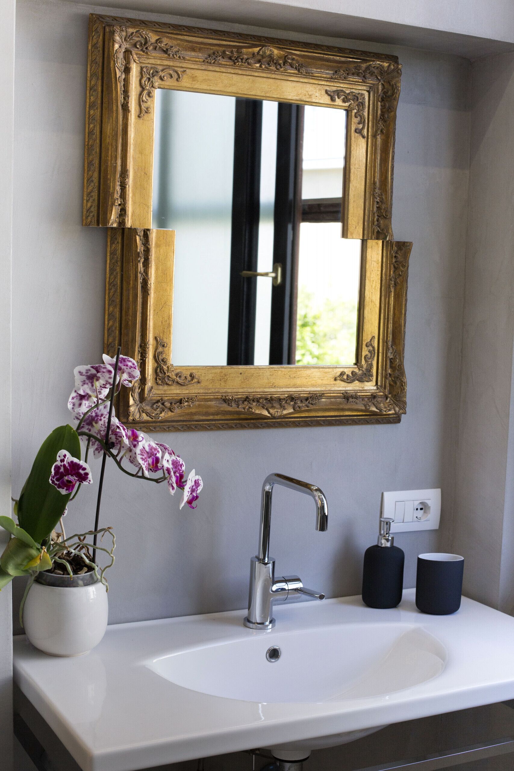 A bathroom with a white sink, a gold-framed mirror, a potted orchid, and two black cups.