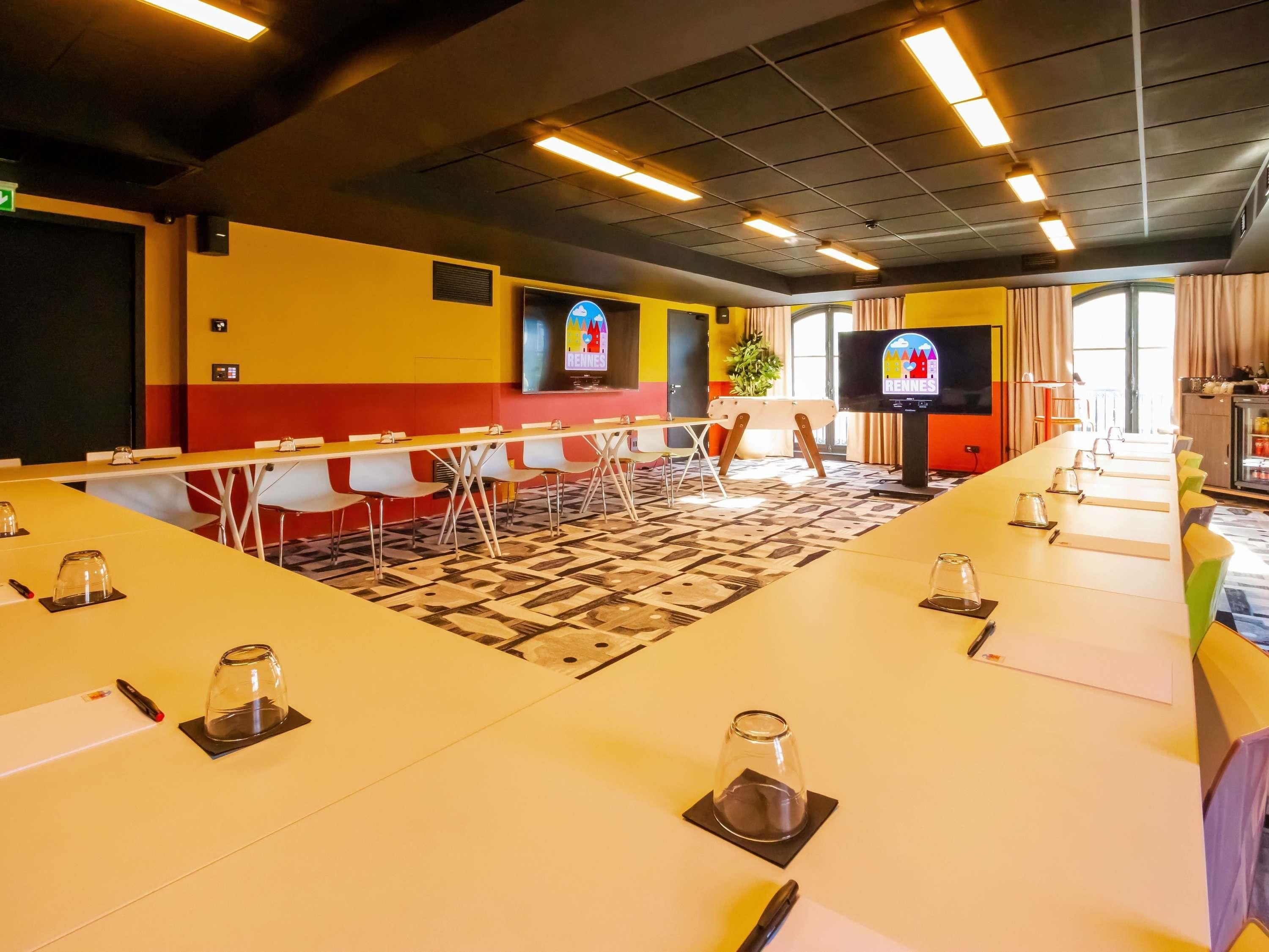 Uma sala de conferências com uma mesa comprida, cadeiras, uma televisão e uma planta.