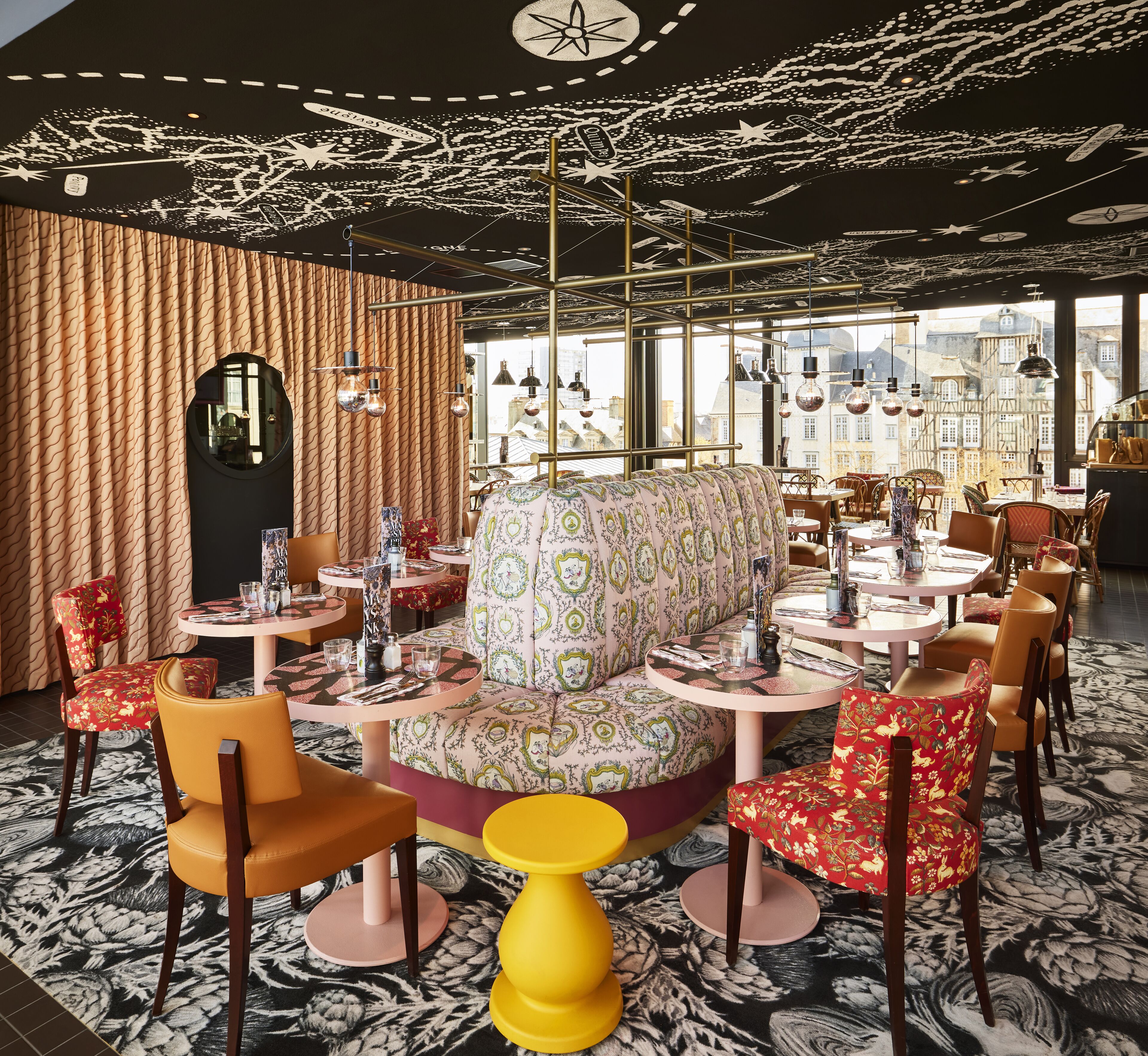 Um restaurante com mesas redondas, estofados estampados e vista da cidade através de grandes janelas.
