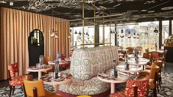 Um restaurante com mesas redondas, estofados estampados e vista da cidade através de grandes janelas.