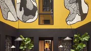 A modern interior with a yellow accent wall, large black and white artwork, and a central elevator. Furnishings include patterned sofas, armchairs, and a round table.