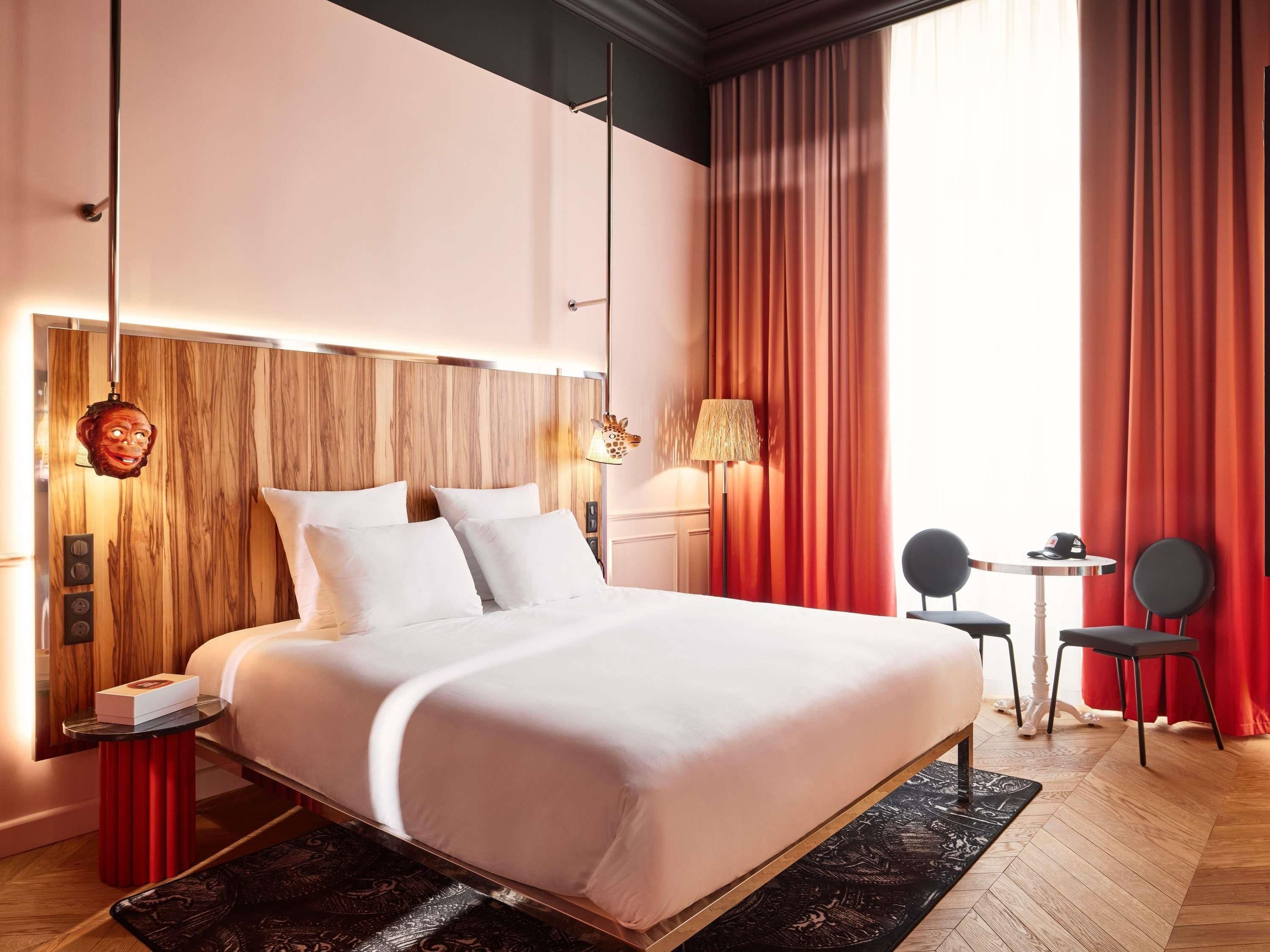 A modern hotel room with a wooden headboard, a bed with white linens, a small table with a chair, and a view of the outdoors through large windows.