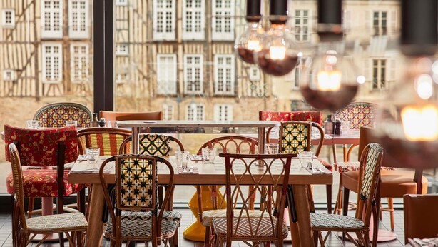 Interior de restaurante com vista para edifícios tradicionais, cadeiras de vime e luminárias pendentes.