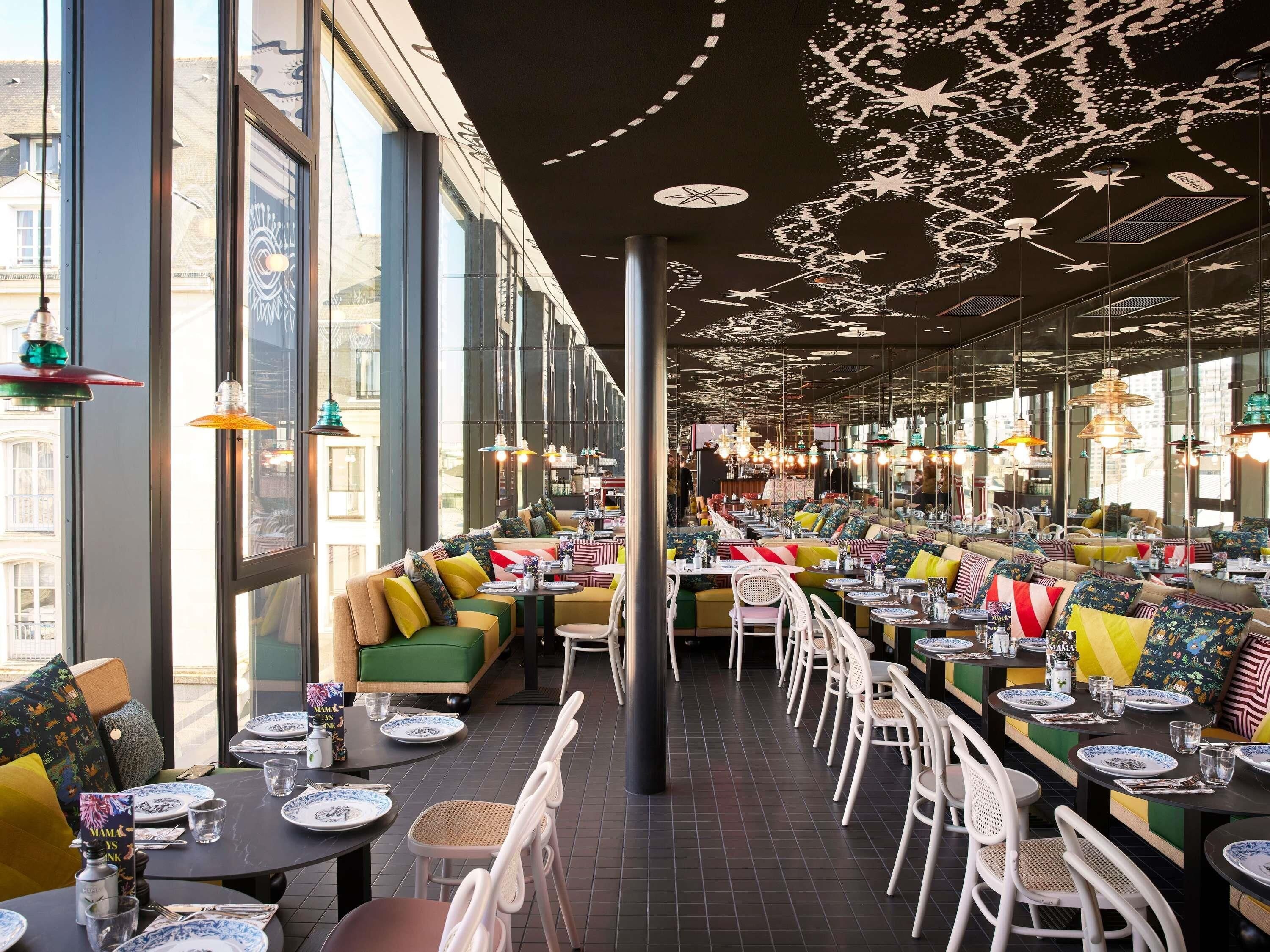 A restaurant with high ceilings, large windows, and colorful seating. Tables are set with plates, glasses, and cutlery.