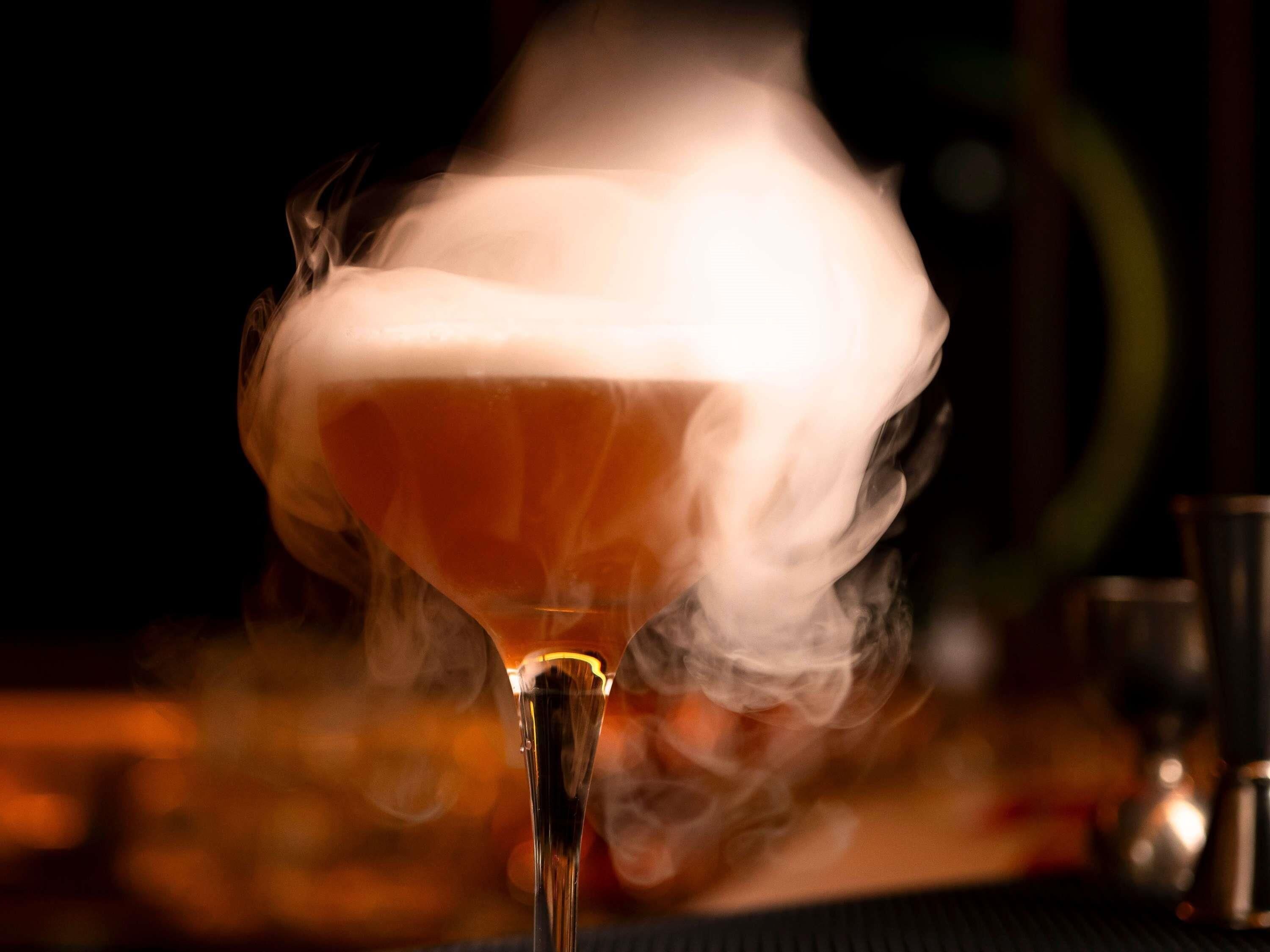 A cocktail with a smoky effect, a clear glass, and a blurred background with indistinct objects.