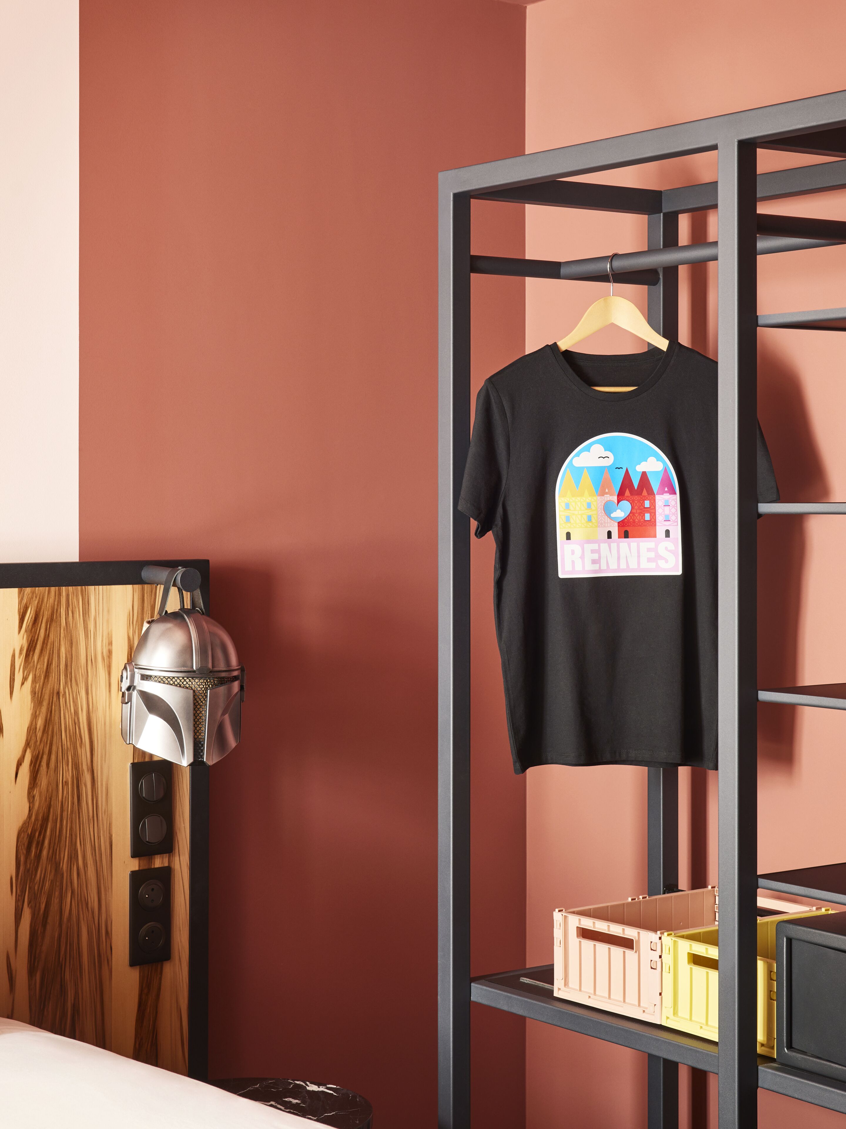 A room with a bed, a wooden headboard, a wall-mounted lamp, a black t-shirt on a hanger, and a metal shelving unit with storage boxes.