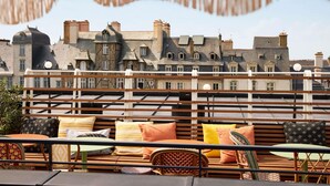 Terraço no telhado com vista para edifícios históricos, móveis de madeira e guarda-corpo.