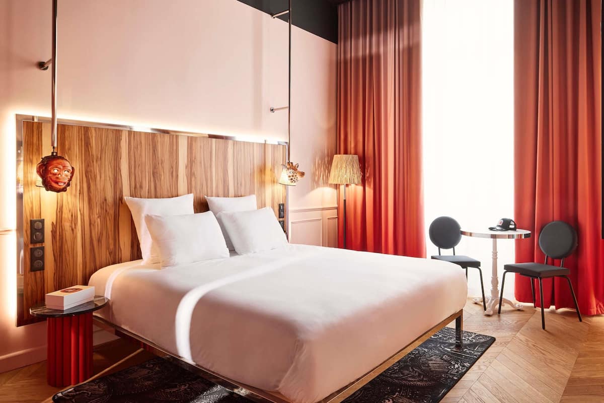 A modern hotel room with a wooden headboard, a bed with white linens, a small table with a chair, and a view of the outdoors through large windows.