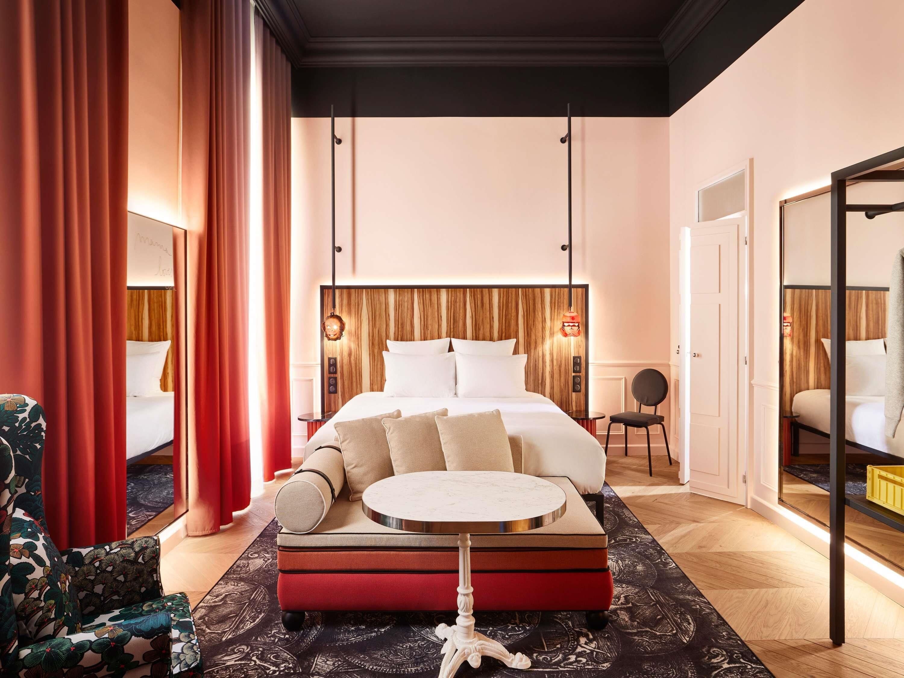 A modern hotel room with a large bed, a round side table, a chair, and a mirror.
