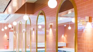 Uma piscina interna com água cristalina, rodeada por paredes de azulejos rosa e espelhos em arco dourados.