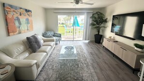 A living room with a sofa, coffee table, TV, and a large window with a view of a balcony. - Beautiful Oceanview Townhouse 2 Bed 2.5 Bath SH 430 (St. Augustine)