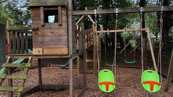 A wooden play structure with a slide and swings, surrounded by mulch and trees.