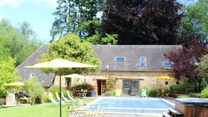 Ein Poolbereich mit Liegestühlen und Sonnenschirmen, ein Steinhaus mit Tür und Fenstern sowie ein Baum mit violetten Blättern.