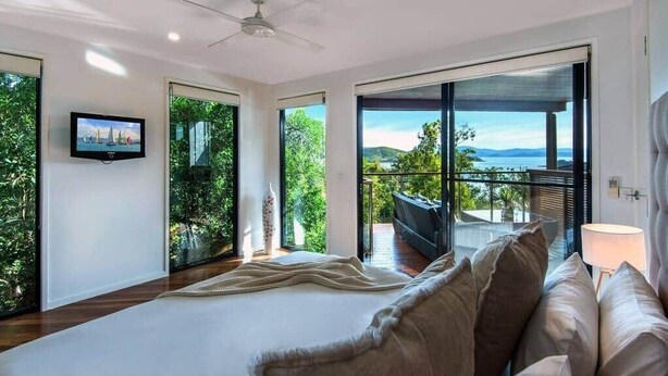 Ein Schlafzimmer mit einem großen Bett, einem Fernseher und einem Balkon mit Blick auf Grünflächen und ein Gewässer.