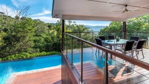 Eine Holzterrasse mit Glasgeländer, einem Pool und einem Tisch mit Stühlen.