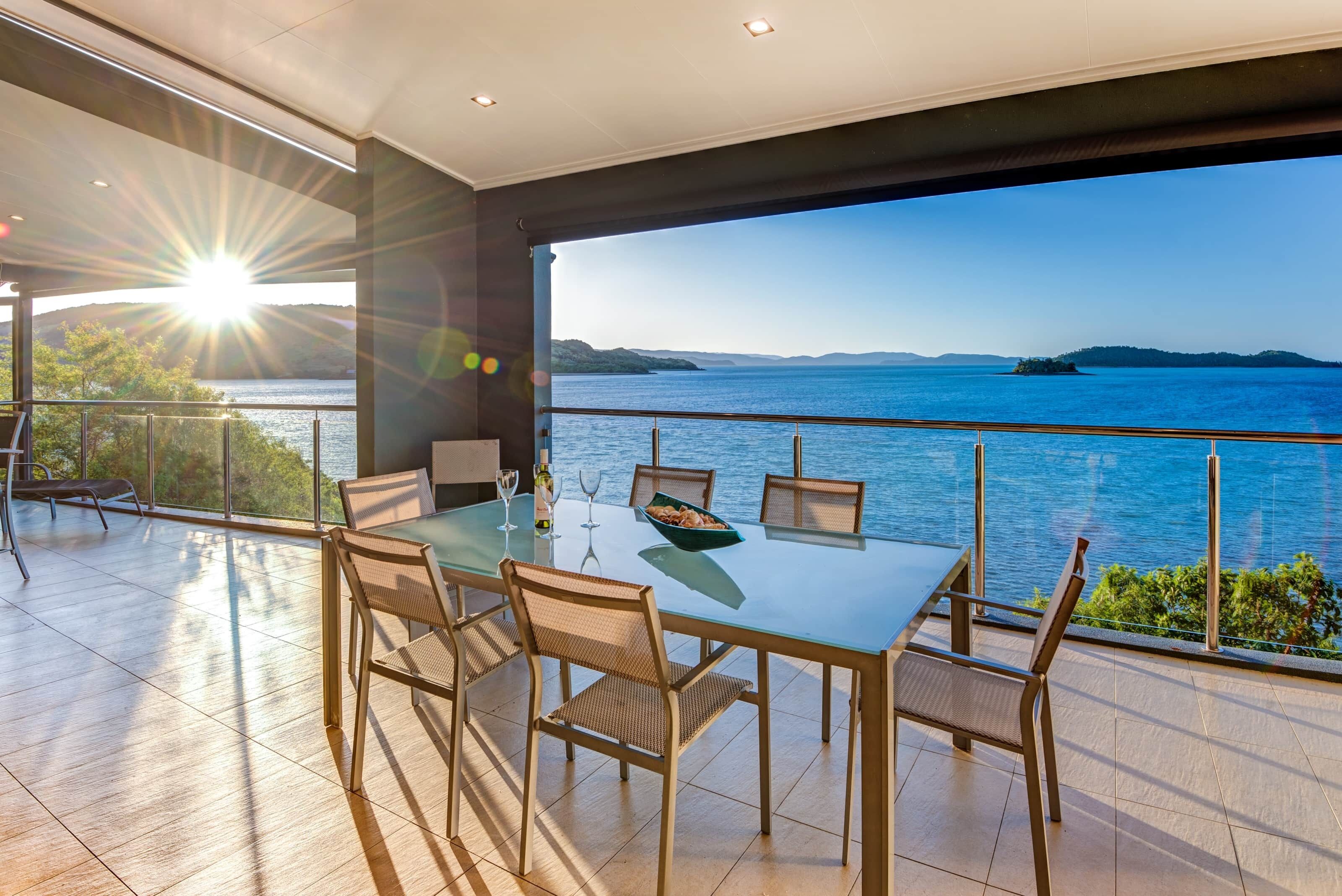 A dining area with a glass table and chairs, overlooking a body of water with a distant island.