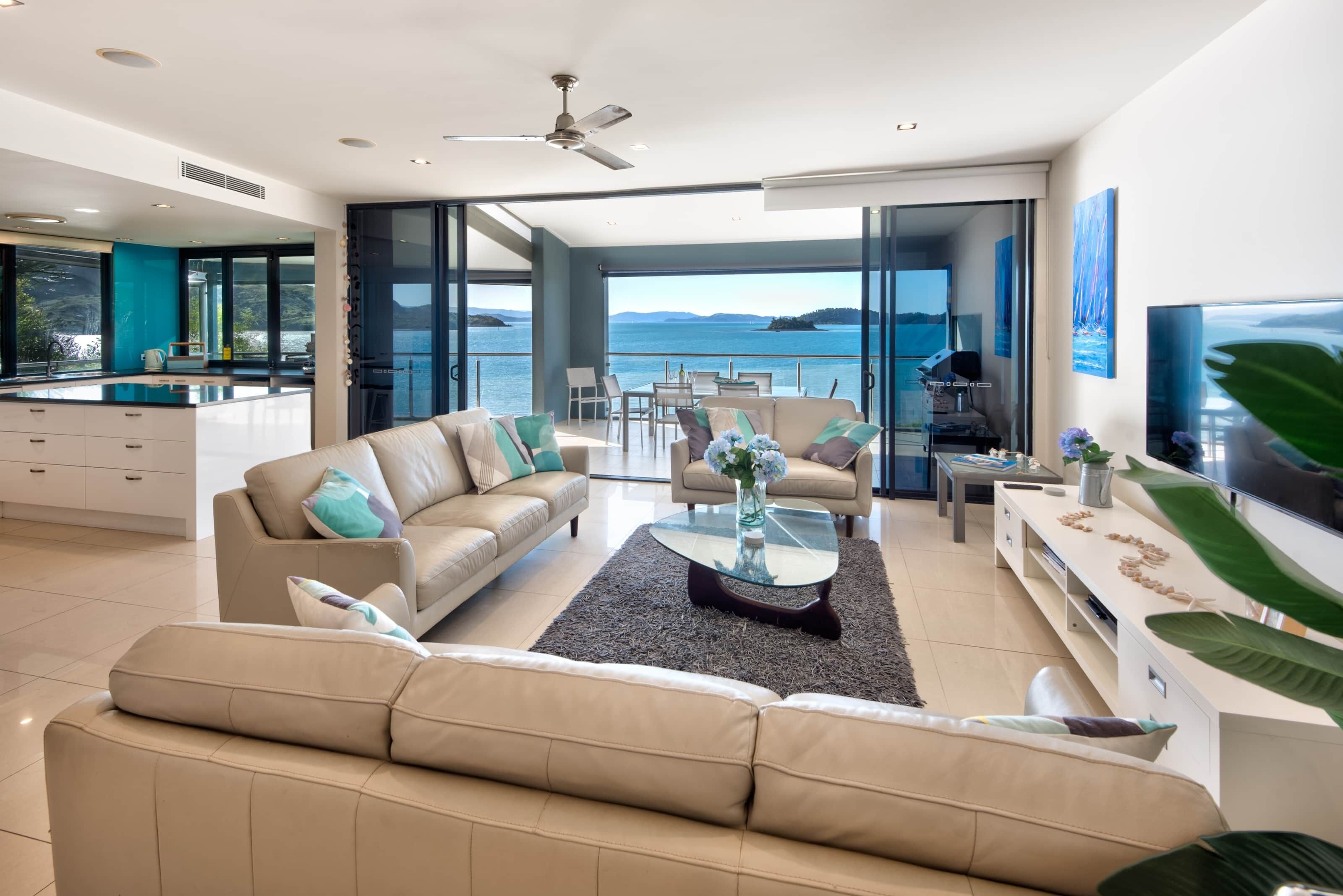 A modern living room with a sofa, coffee table, and TV. There is a balcony with a view of the sea.