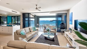 A modern living room with a sofa, coffee table, and TV. There is a balcony with a view of the sea.