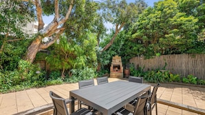 A patio with a table and chairs, surrounded by lush greenery and a wooden fence. - Esplanade Premium 33 Steps to Beach (Torquay)