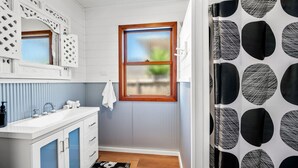 A bathroom with a white vanity, a window with a wooden frame, and a patterned curtain. - Esplanade Premium 33 Steps to Beach (Torquay)