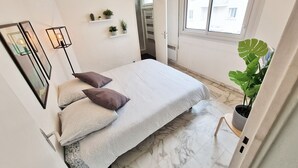 Un dormitorio con cama, almohadas, una planta en maceta, una ventana y una puerta que da a un baño.