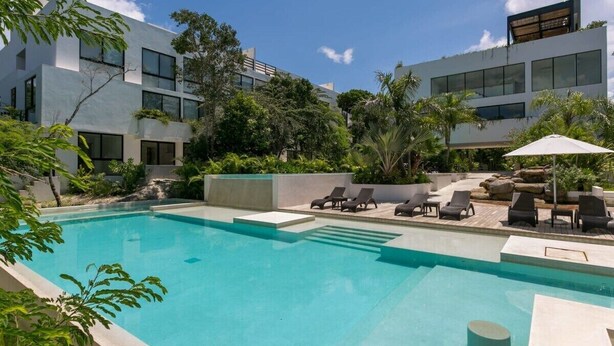 A modern pool area with a clear blue pool, lounge chairs, and a building with large windows and greenery. - 2BR Condo Beautiful Common Areas, Sleeps 6! (Akumal)