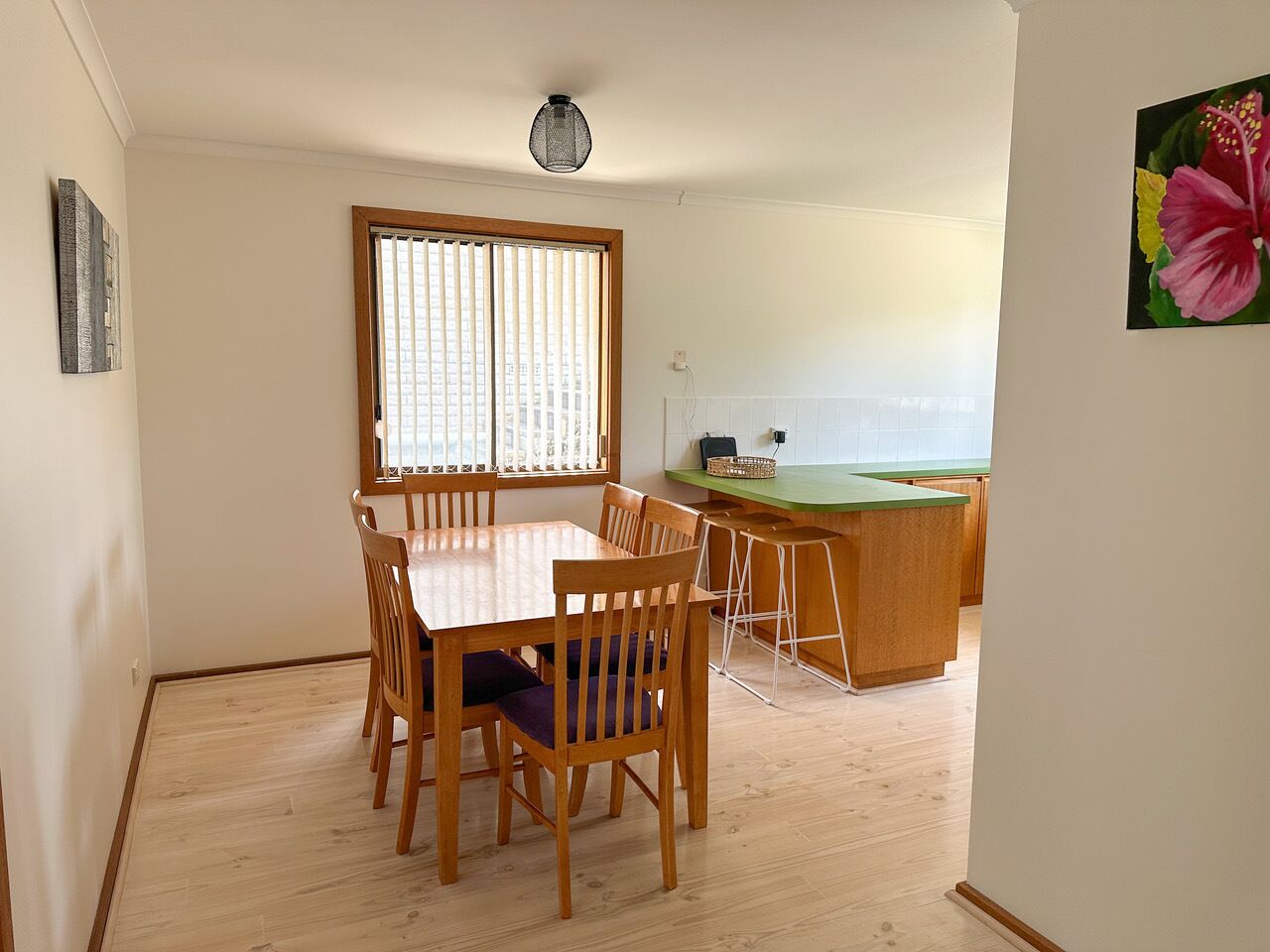 A dining area with a wooden table and chairs, a kitchen counter, and a window with blinds.