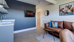 A modern living room with a brown leather sofa, a mounted TV displaying 'NETFLIX', a painting on the wall, and a herringbone wood floor.