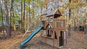 A wooden playhouse with a slide in a wooded area. - Pet-Friendly Playset Retreat for Families (Broken Bow)