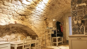Una habitación rústica con paredes de piedra, una mesa de comedor con sillas, una mesita auxiliar con una silla, y una zona de cocina con fregadero y salpicadero de azulejos.