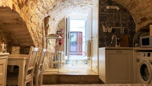 Habitación rústica con paredes de piedra, cama, mesa, sillas, lavadora y una zona de cocina con microondas.