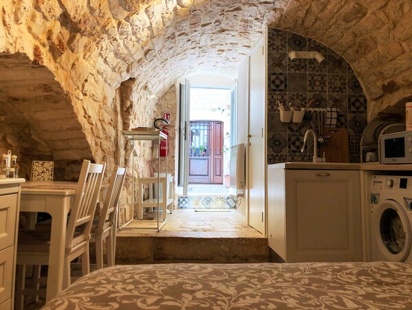 Habitación rústica con paredes de piedra, cama, mesa, sillas, lavadora y una zona de cocina con microondas.