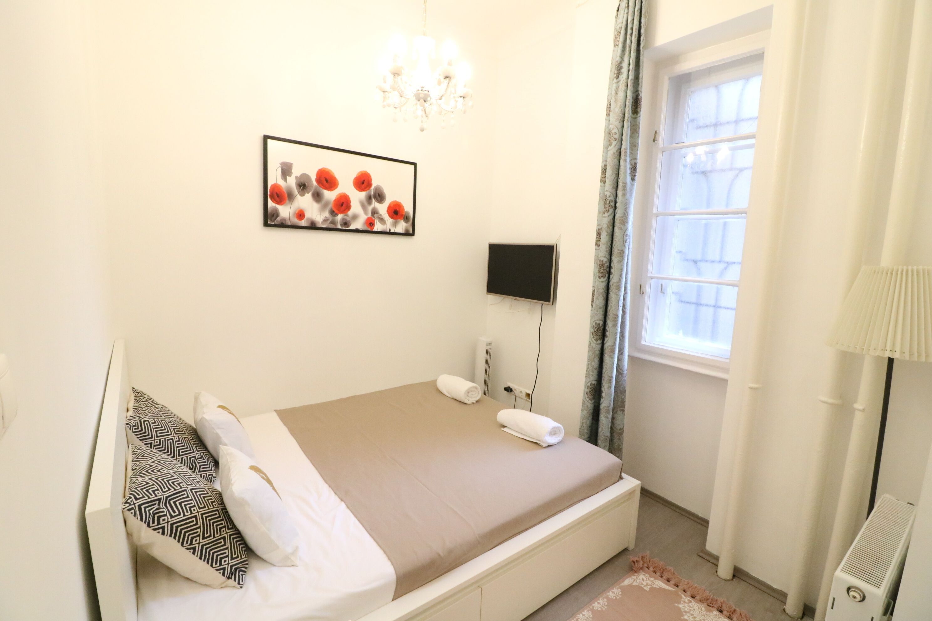 A neatly arranged bedroom with a bed, a wall-mounted TV, a window with curtains, and a chandelier.