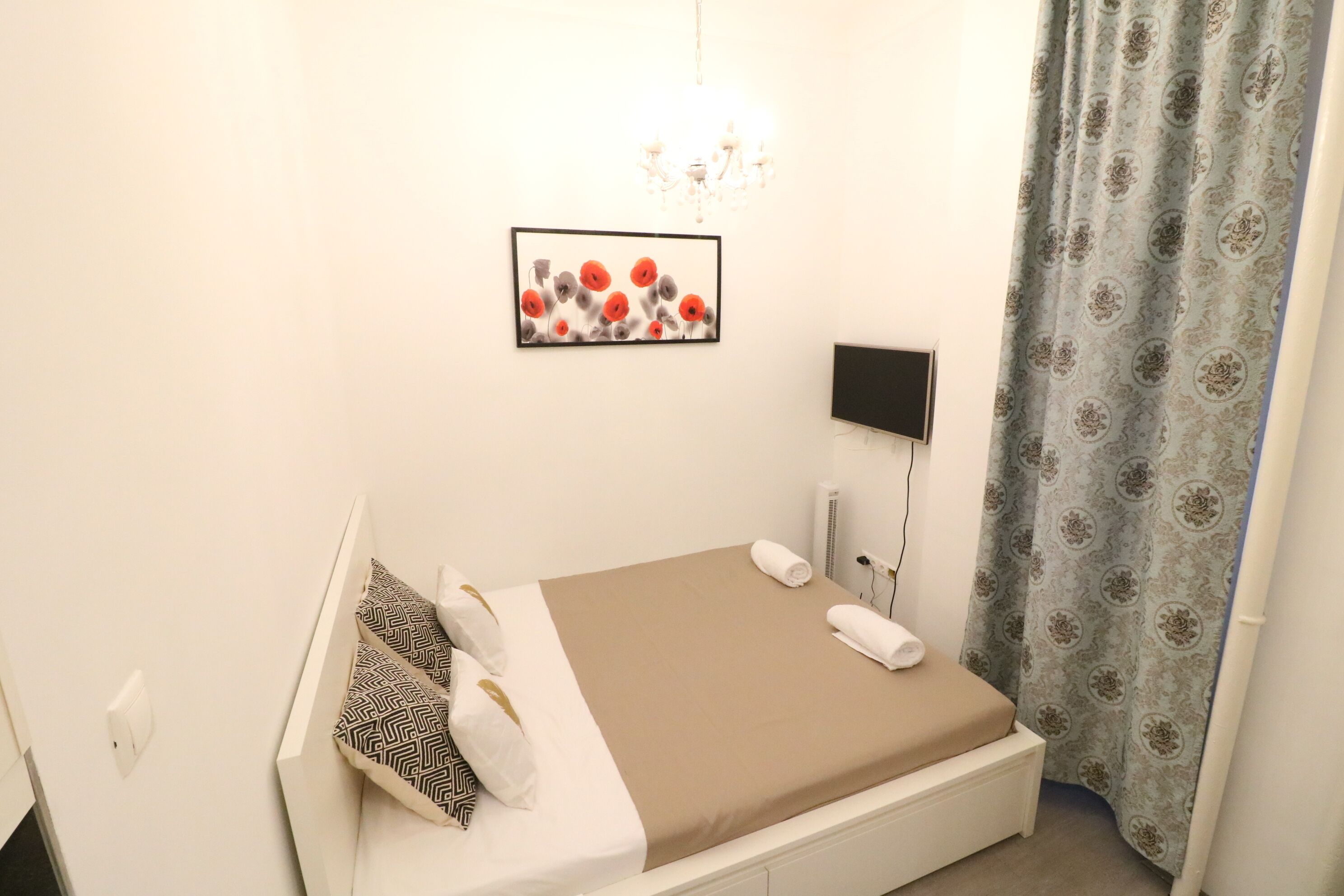 Een netjes opgemaakt bed met een bruine matras en beddengoed met patronen, een tv aan de muur, een ingelijste afbeelding en een gordijn met een patroon.