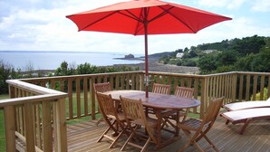 Eine Holzterrasse mit einem runden Tisch, Stühlen, einem roten Sonnenschirm und Blick aufs Meer.