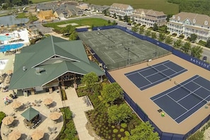 A tennis court surrounded by a covered area with outdoor seating and a pool.