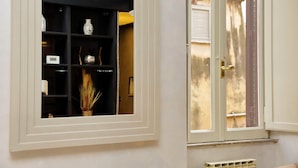 Una habitación con una puerta blanca, una ventana que da al exterior y una estantería con varios objetos, incluyendo un florero y una planta pequeña.