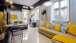 Una sala moderna con sofá, mesa de comedor y sillas. Hay un ventanal con cortinas, un radiador y una planta sobre la mesa.