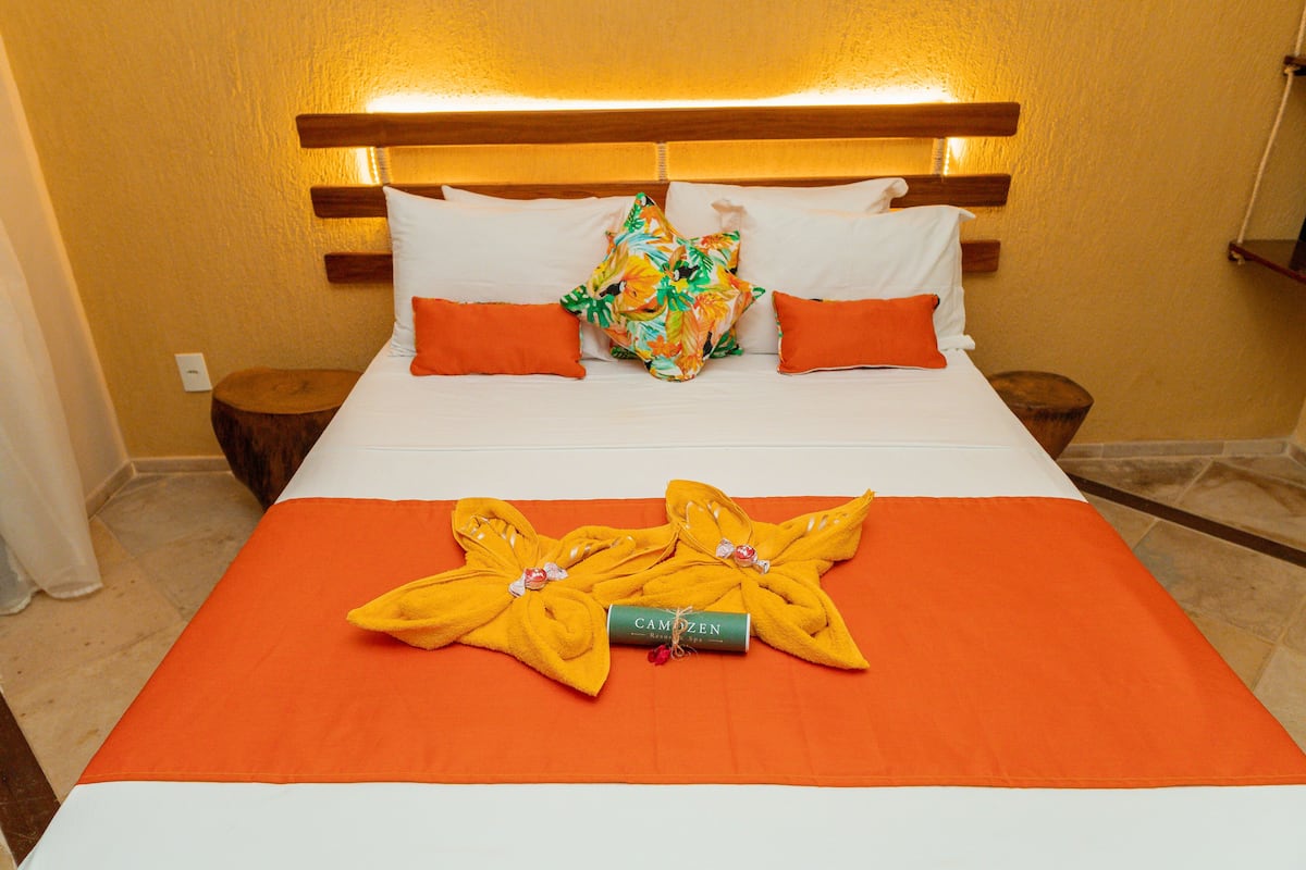 Una cama bien hecha con una colcha naranja, almohadas blancas y una almohada floral de colores. Hay dos mesitas de noche de madera con una pequeña mesa en cada una.